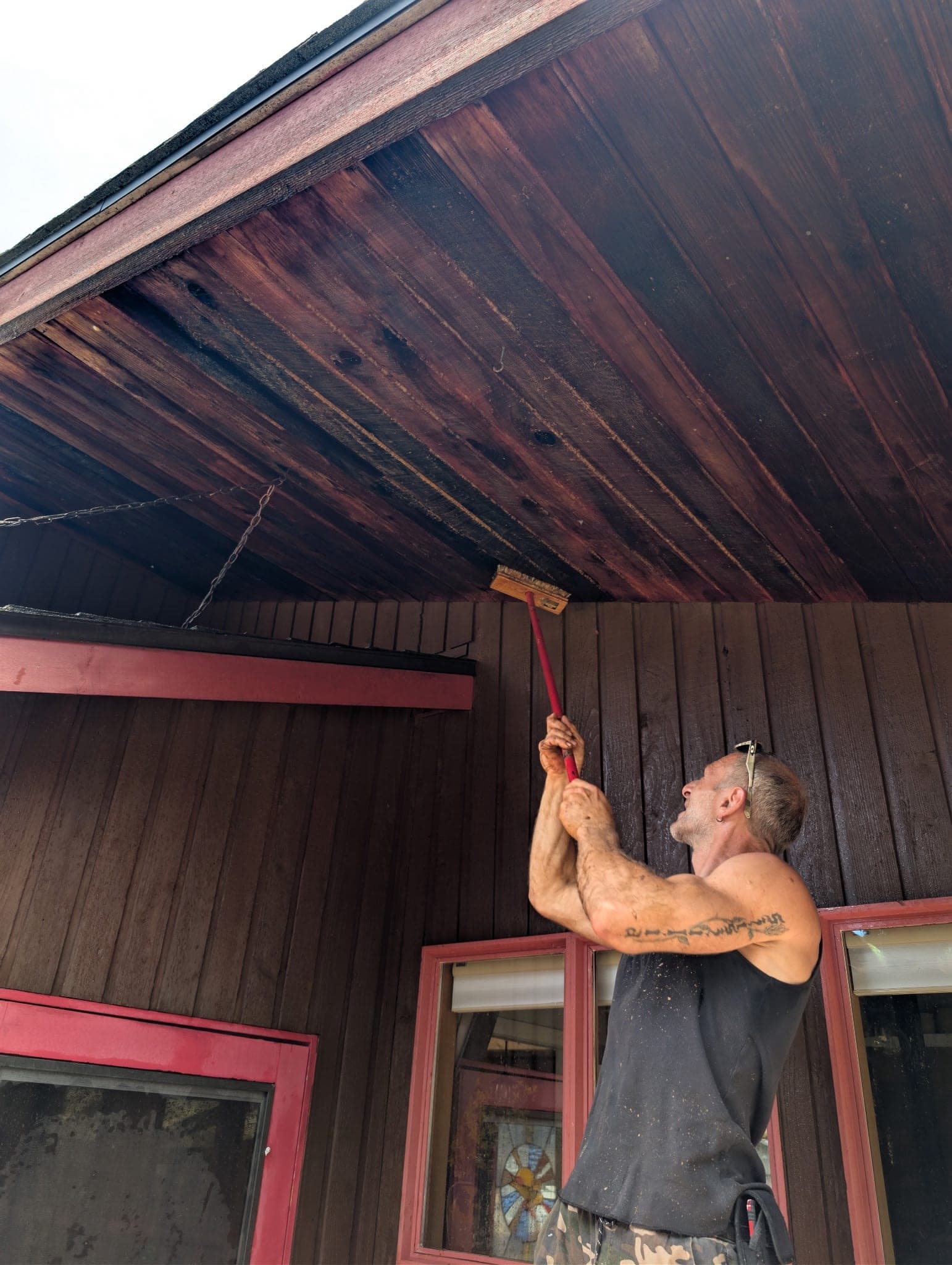 A crew member laying dark stain onto the cedar soffit with a brush on a red extension pole, maroon window trim below