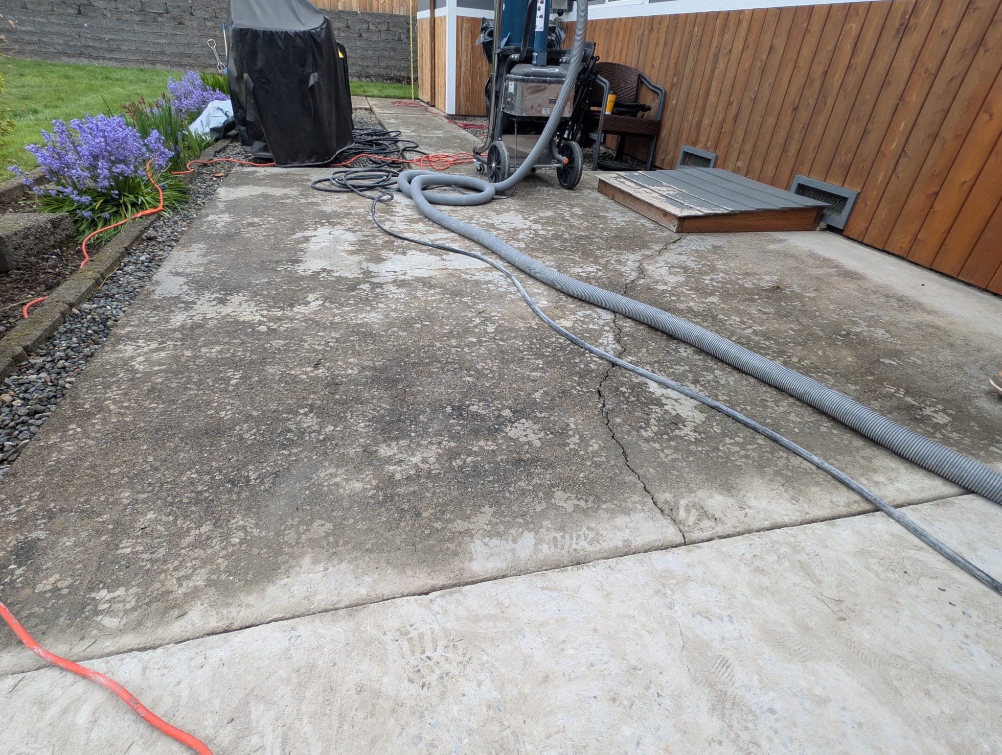 A covered concrete patio blotched grey-green with lichen staining, a crack in the slab and grinder hoses staged on it