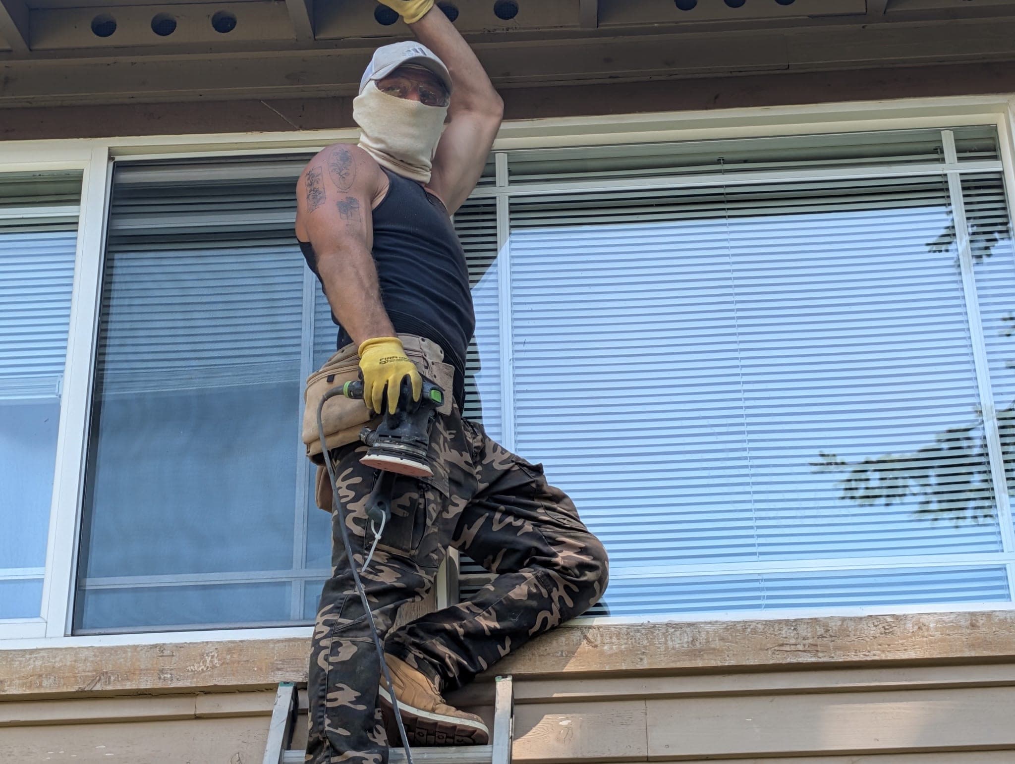 A Painting with Purpose crew member on a ladder, sanding the fascia under the eave with an orbital sander