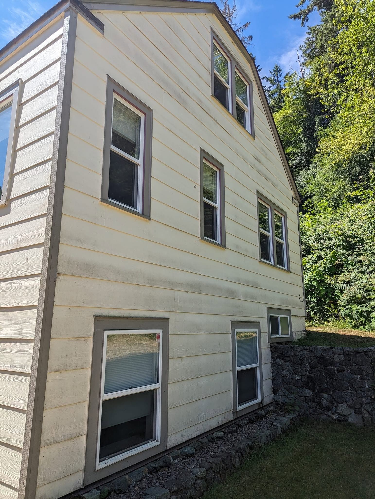 A gambrel-roofed house in tired cream siding with tan trim, streaked from weathering