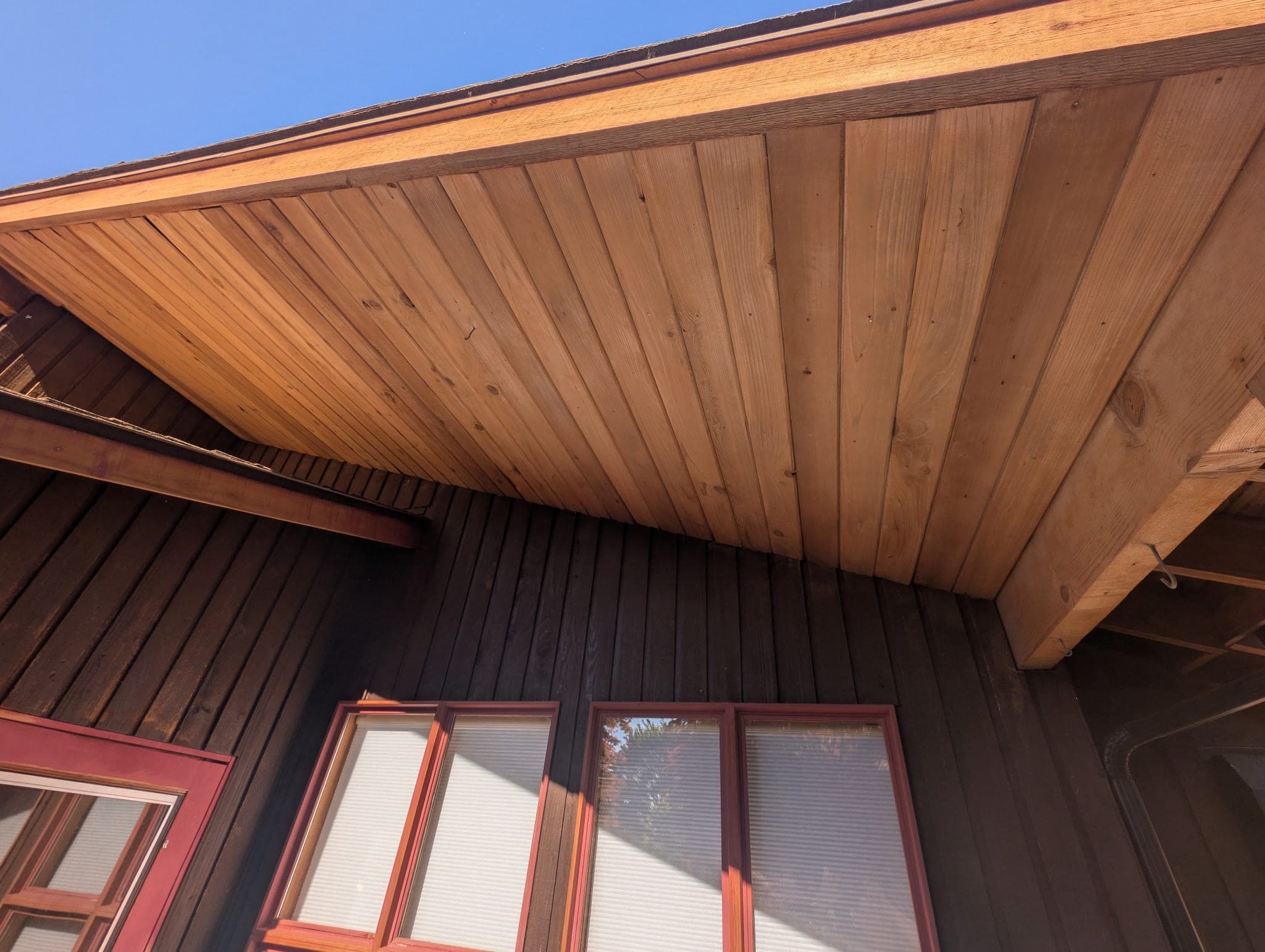 Cedar soffit and fascia sanded back to bare pale wood, above dark stained board and batten siding and maroon window trim
