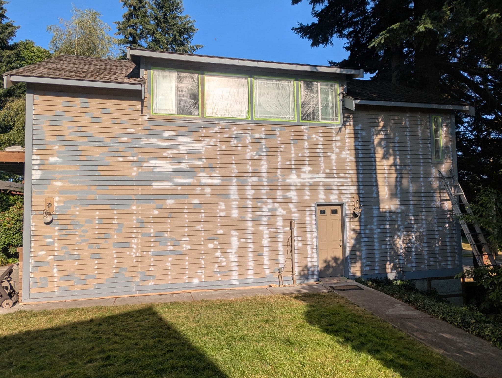 A two-story elevation covered in pale spot-primer patches over failed paint, windows masked and taped out in green
