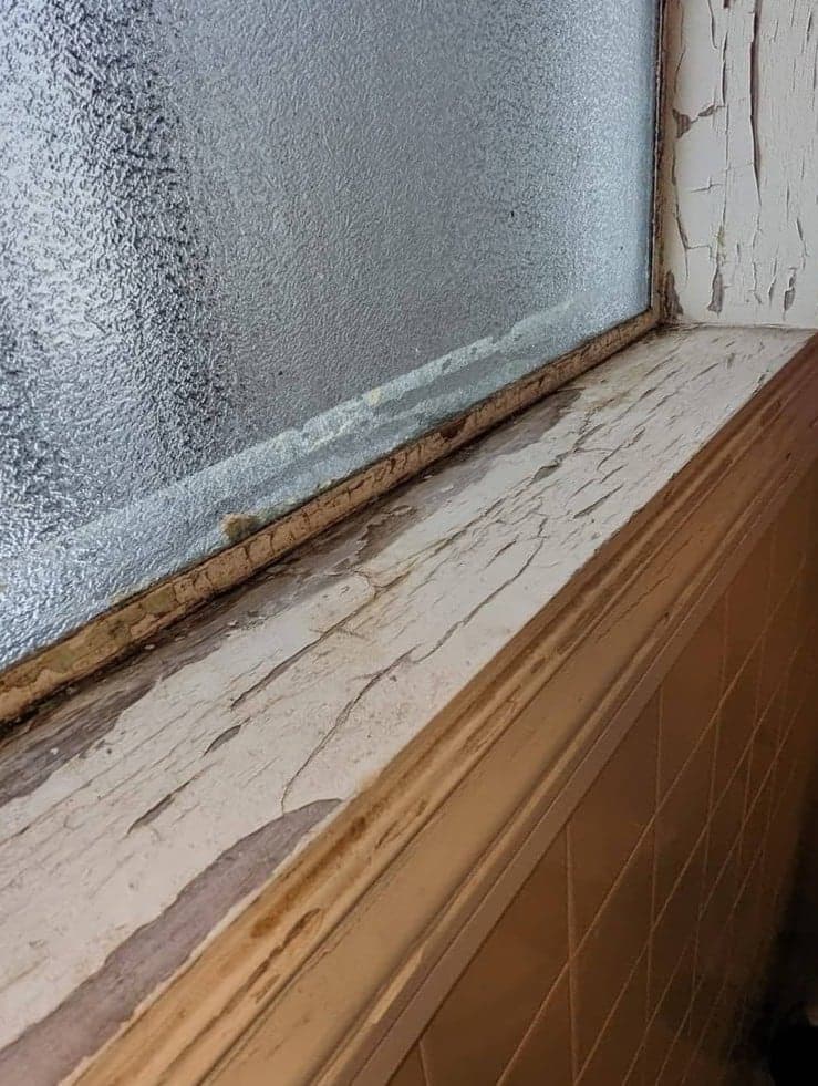 An interior window sill with old paint cracked and lifting away and staining in the corner of the frame