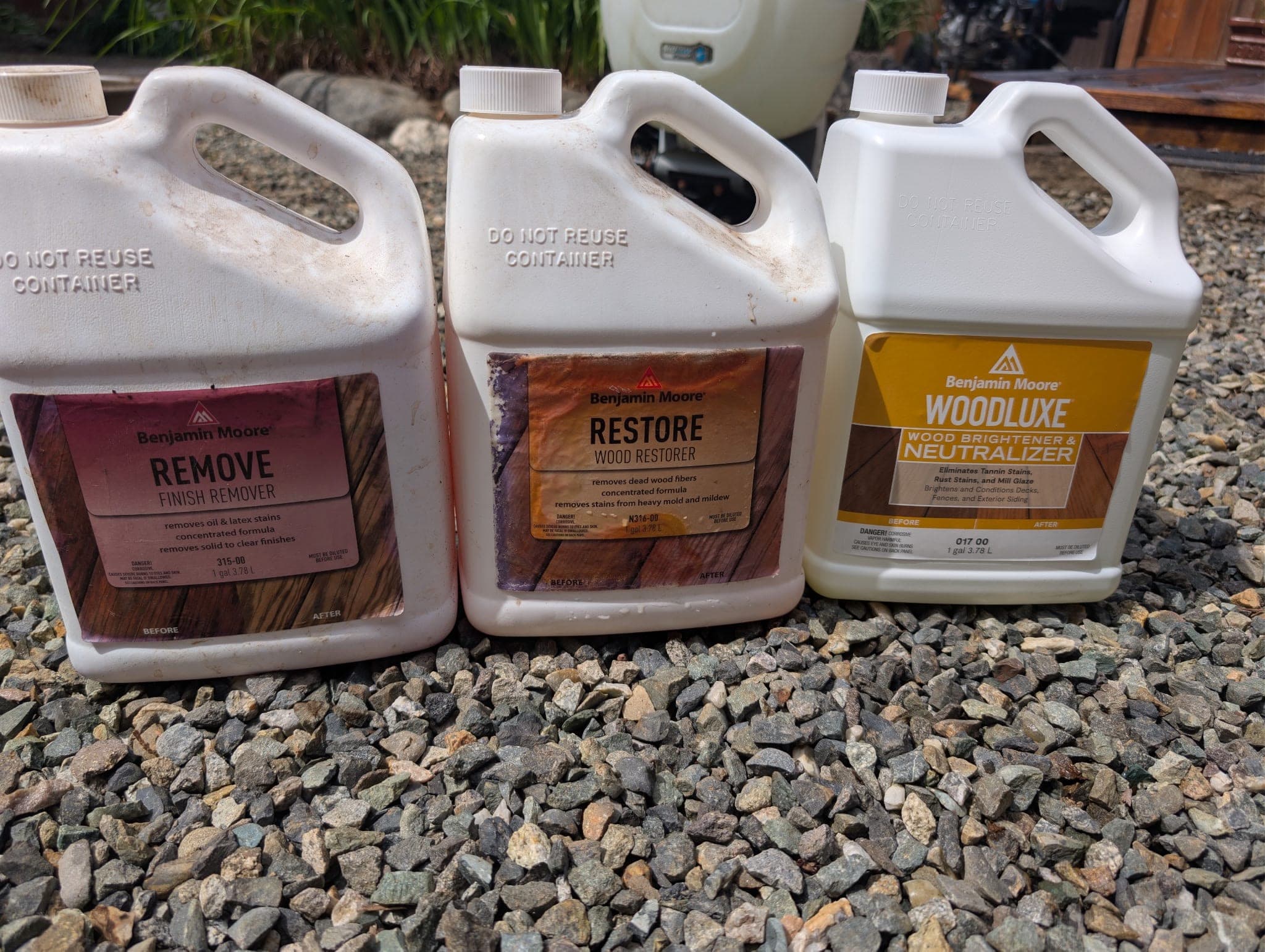 Three Benjamin Moore jugs on gravel, a finish remover, a wood restorer and a wood brightener and neutralizer